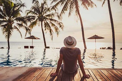 Mann mit Hut sitzt am Pool direkt am Strand unter Palmen. 